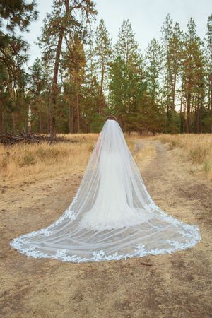 Long cathedral veil with lace trim laid out on dirt path surrounded by pine trees.