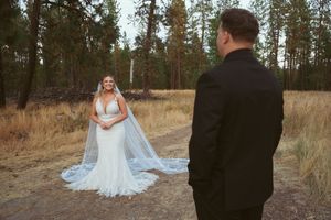 Wedding couple poses together in a rustic outdoor field with tall pine trees in the background.