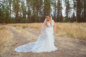 A bride in white lace gown poses on dirt path with flowing veil in natural woodland setting.