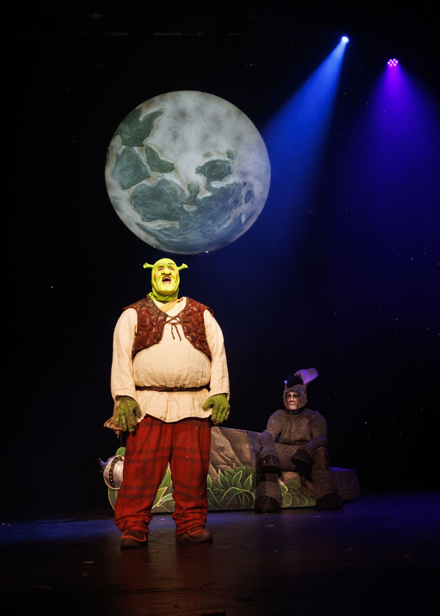 A theatrical stage performance of Shrek with moody lighting and a large moon backdrop showing different sequential poses.
