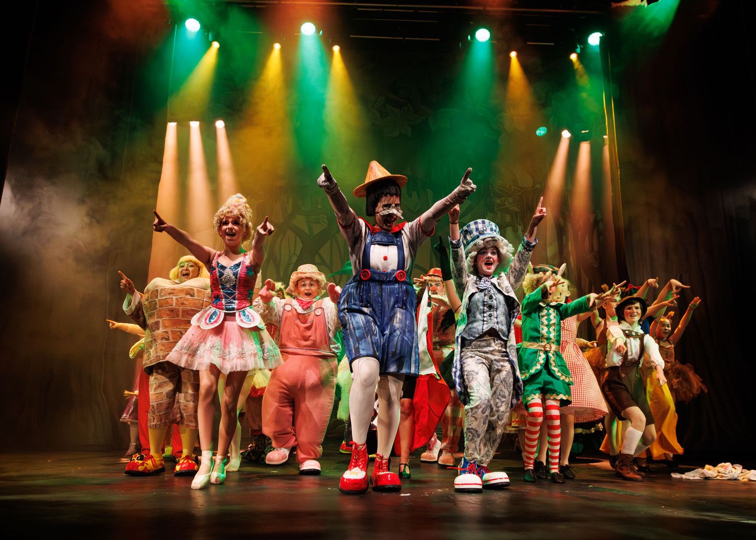 Costumed performers in colorful outfits dance on stage under dramatic green and yellow theatrical lighting during a show.