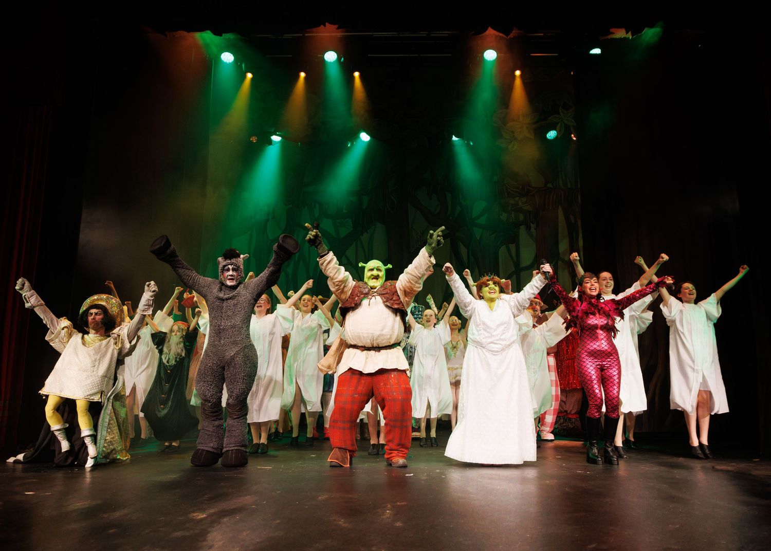 Performers in holiday costumes dance joyfully on stage under dramatic green and red theatrical lighting during a festive show.