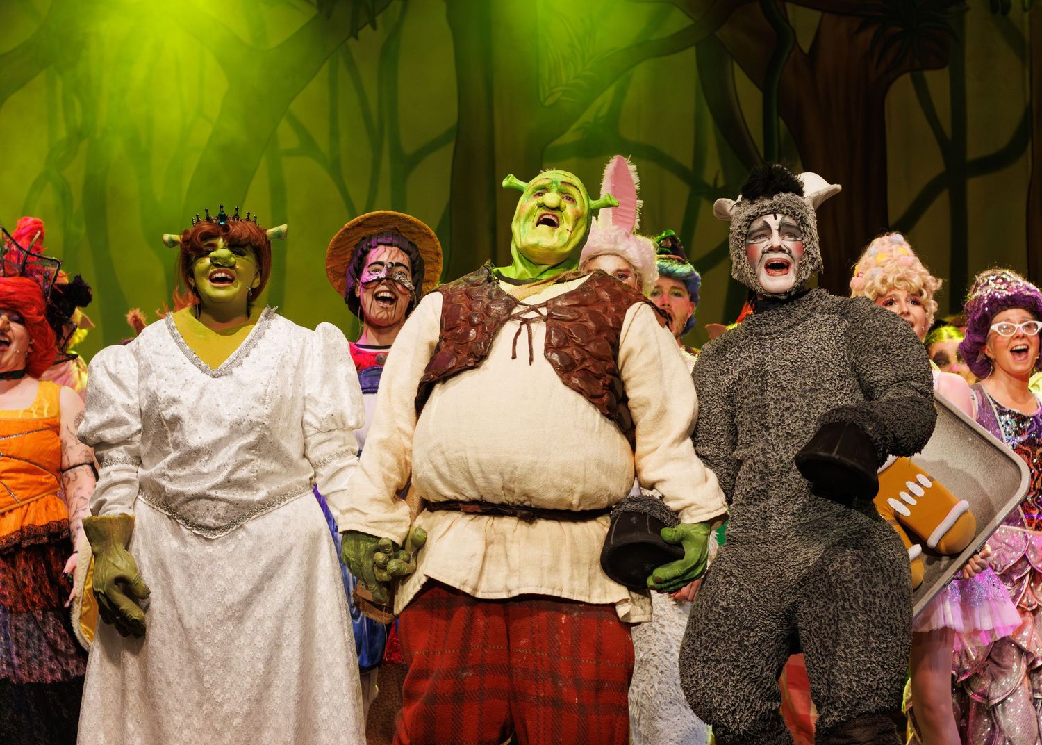 Cast members in costumes performing in a stage production of Shrek the Musical with green forest backdrop.