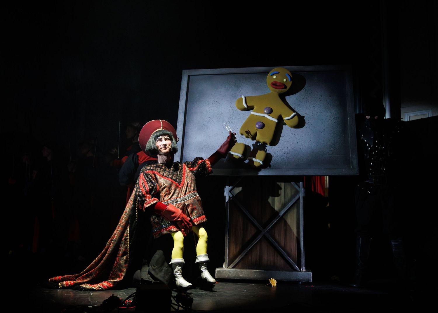 A puppet in a red cloak sits on a chair watching a glowing screen displaying a yellow figure in motion.