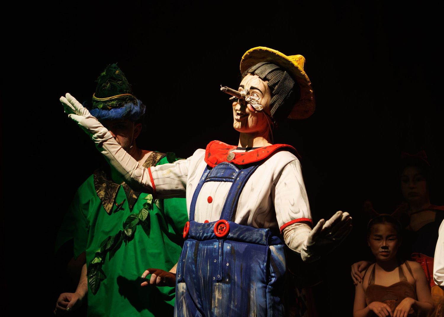 A colorfully dressed performer in puppet-like makeup and costume gestures dramatically on a darkened stage during a show.