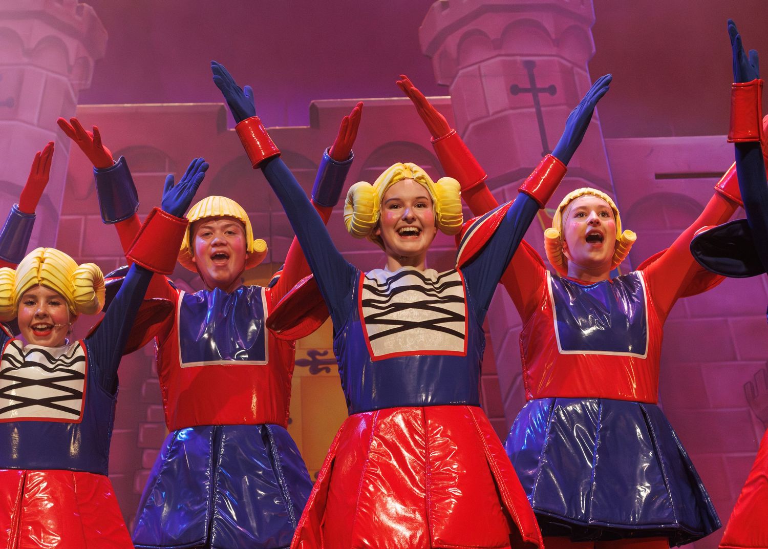 Excited performers in red and blue costumes raise their arms in joyful celebration on a vibrant stage.