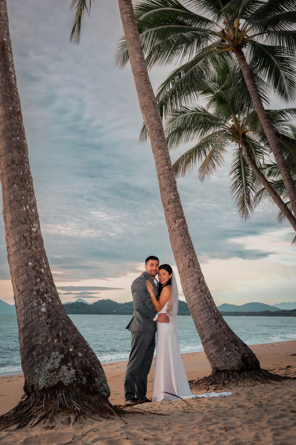 A romantic beach scene with palm trees framing a couple on a tropical shore at sunset with mountains in the distance.
