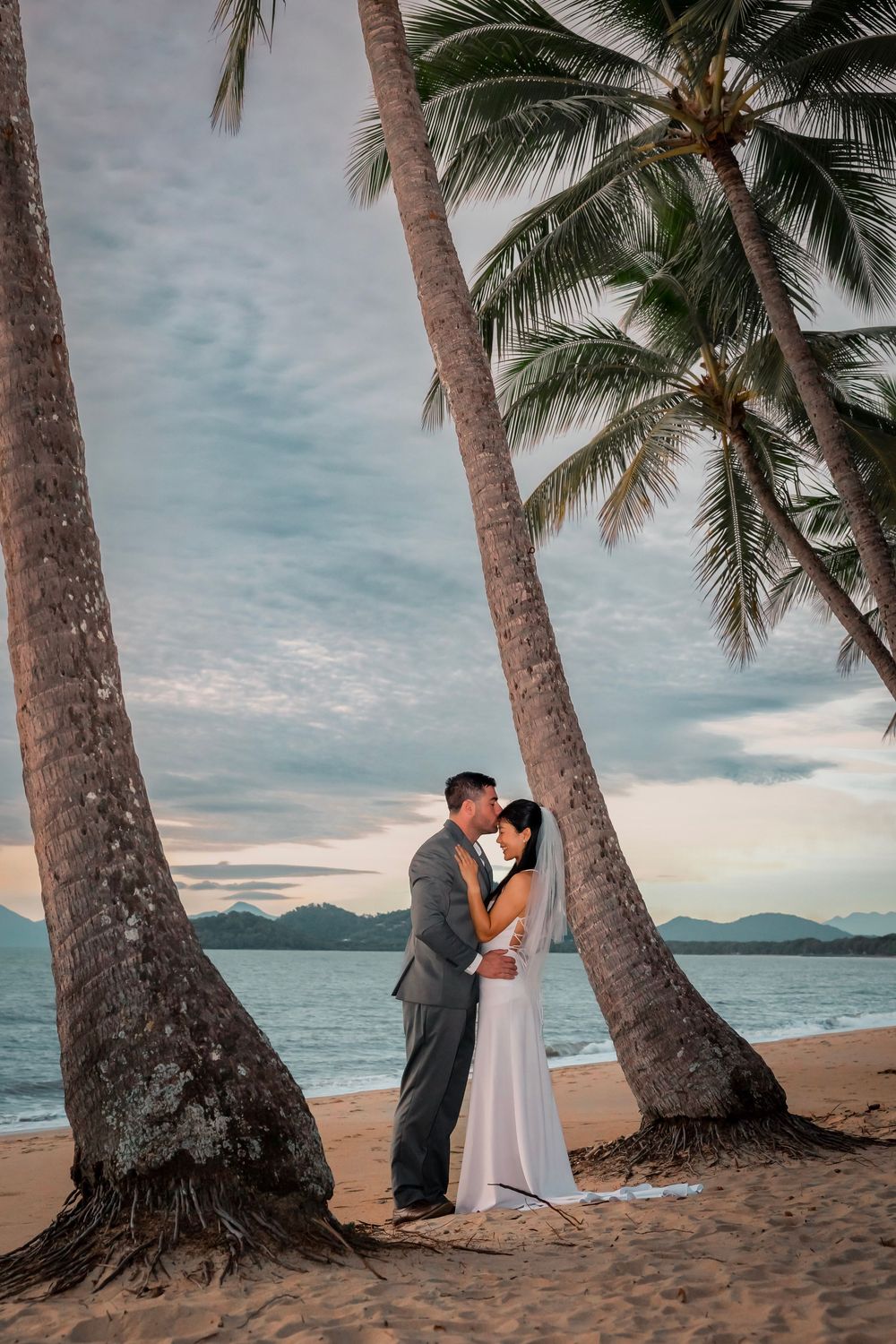 A couple shares an intimate moment beneath palm trees on a tropical beach at sunset with ocean views.