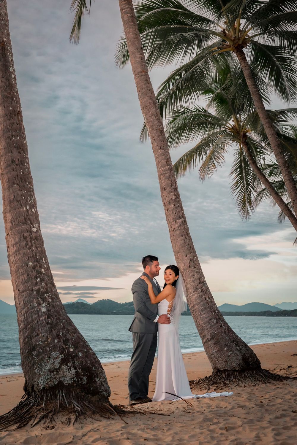 A romantic beach wedding photo at sunset with palm trees framing the couple on a tropical shore.