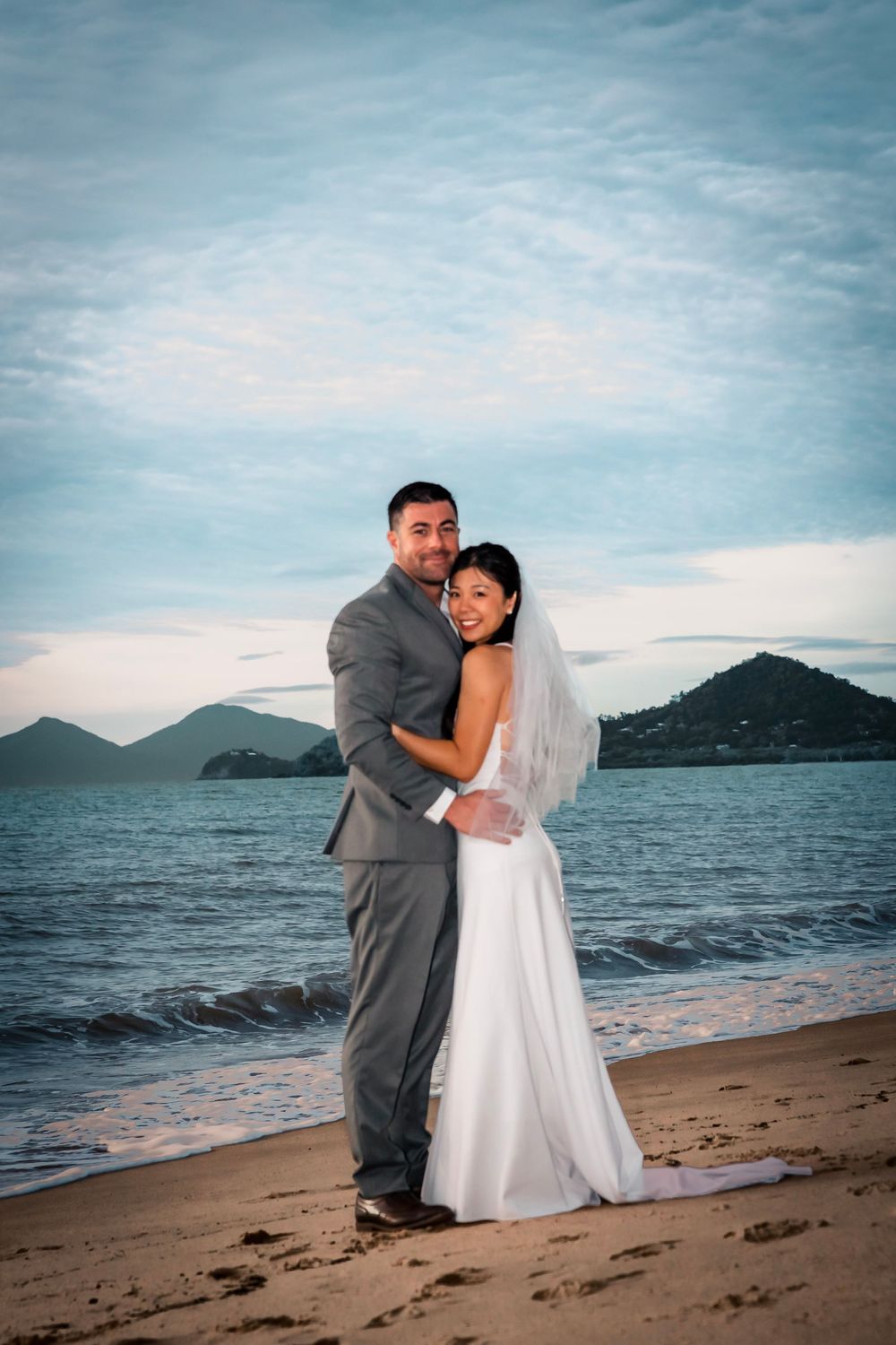 A newlywed couple embraces on a beach at sunset with mountains and ocean in the background.