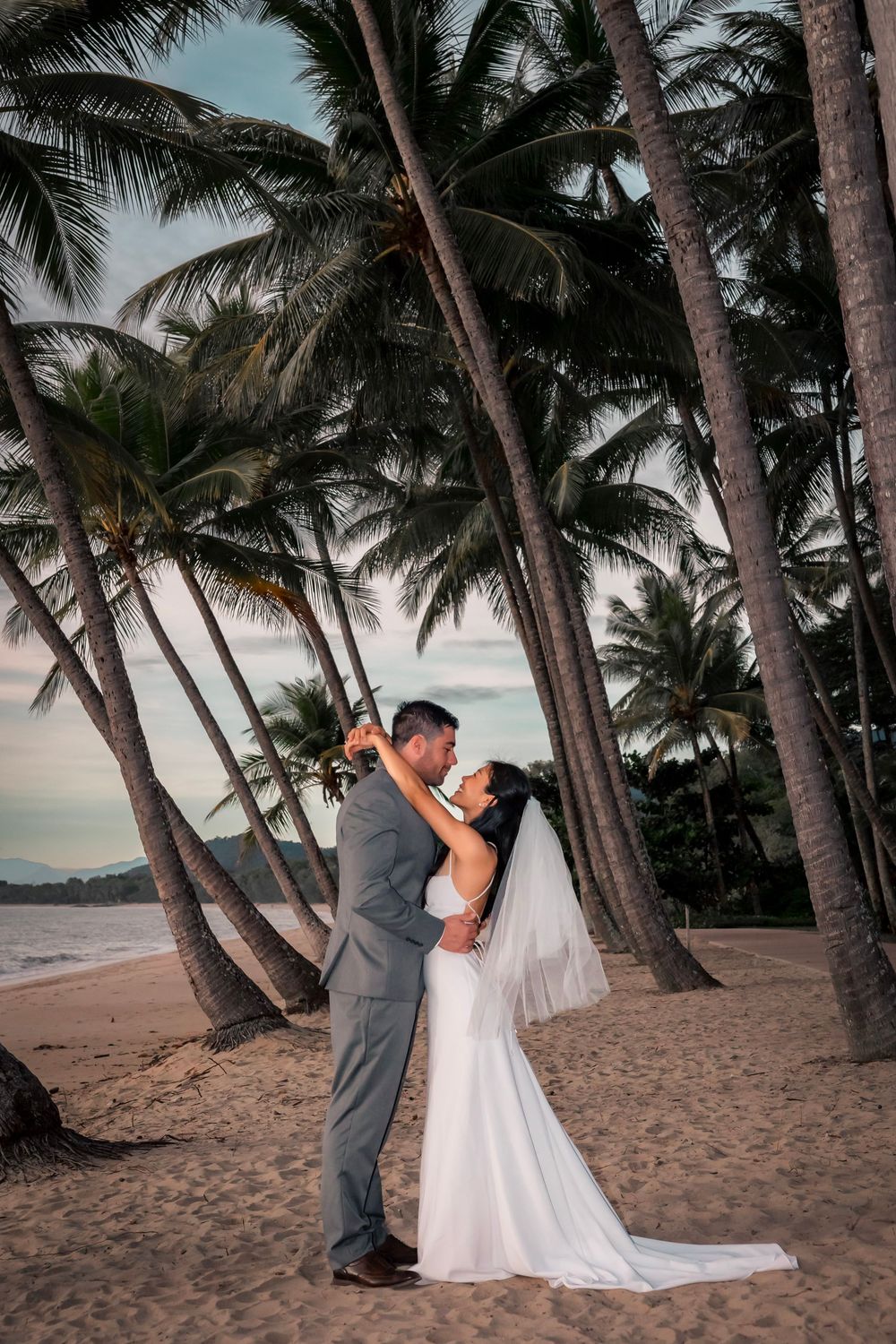 A couple embraces on a tropical beach at sunset surrounded by towering palm trees in Hawaii.