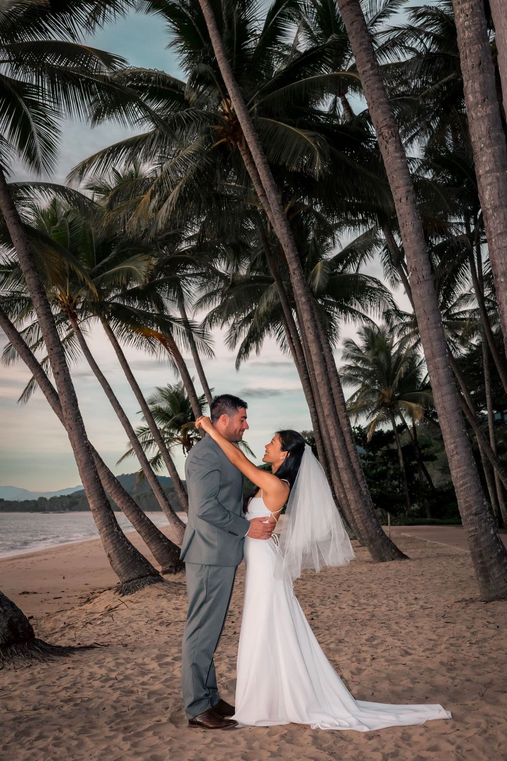Newlywed couple shares romantic moment on a tropical beach at sunset surrounded by towering palm trees.