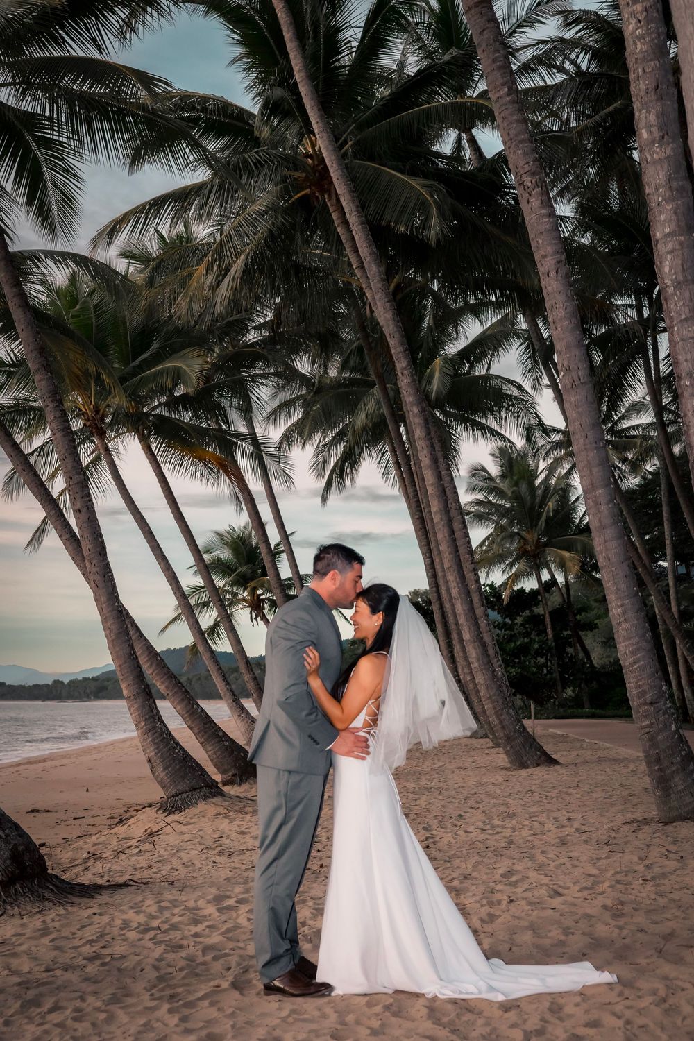 A couple embraces on a tropical beach at sunset surrounded by leaning palm trees in Hawaii.