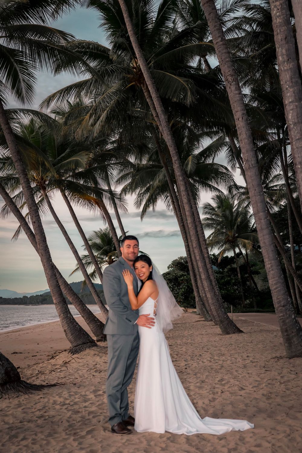 A romantic couple poses on a tropical beach at sunset surrounded by tall palm trees on the sandy shore.
