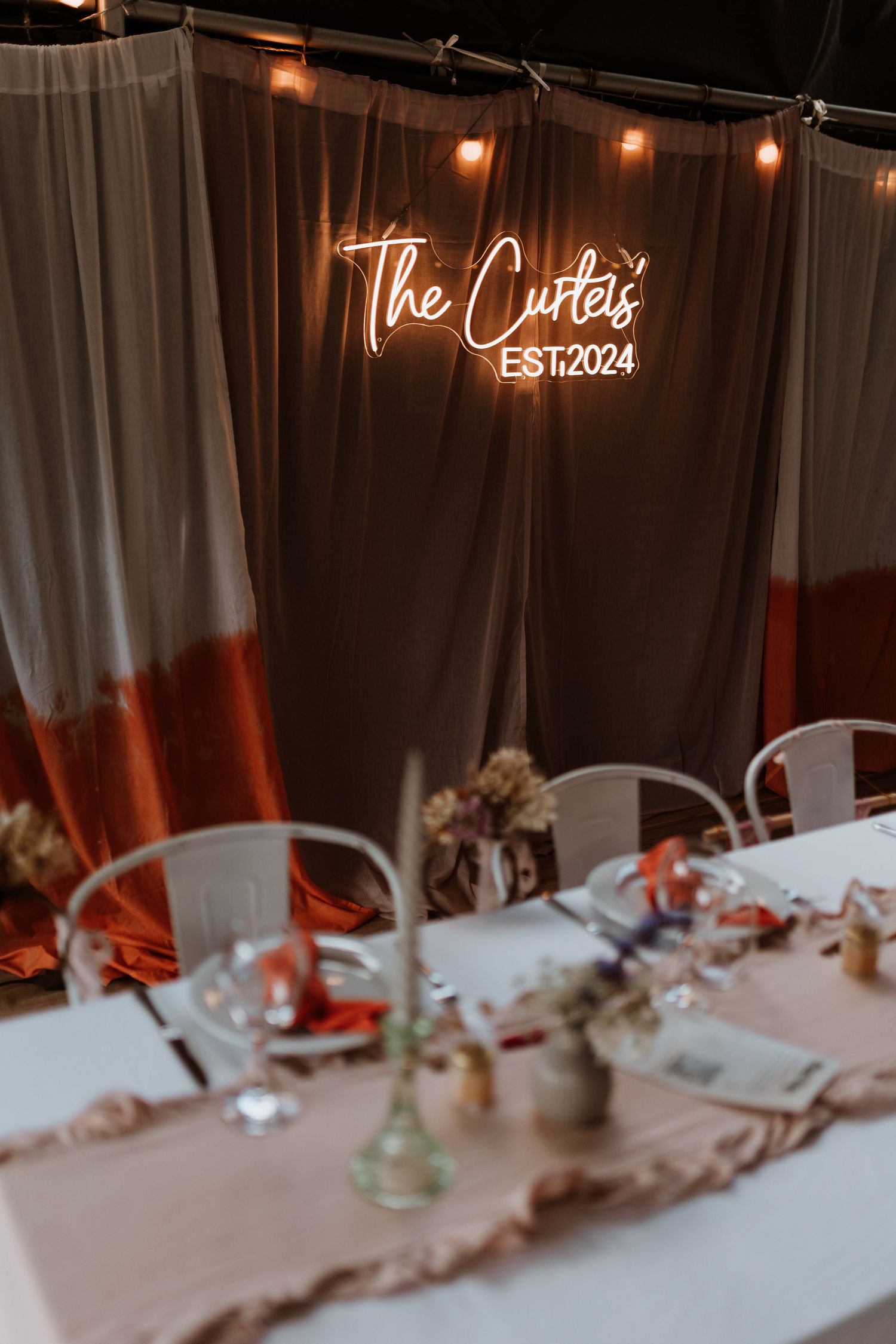 Elegant wedding table setting with neon sign reading 'The Curtis EST 2024' against dark curtains and string lights.