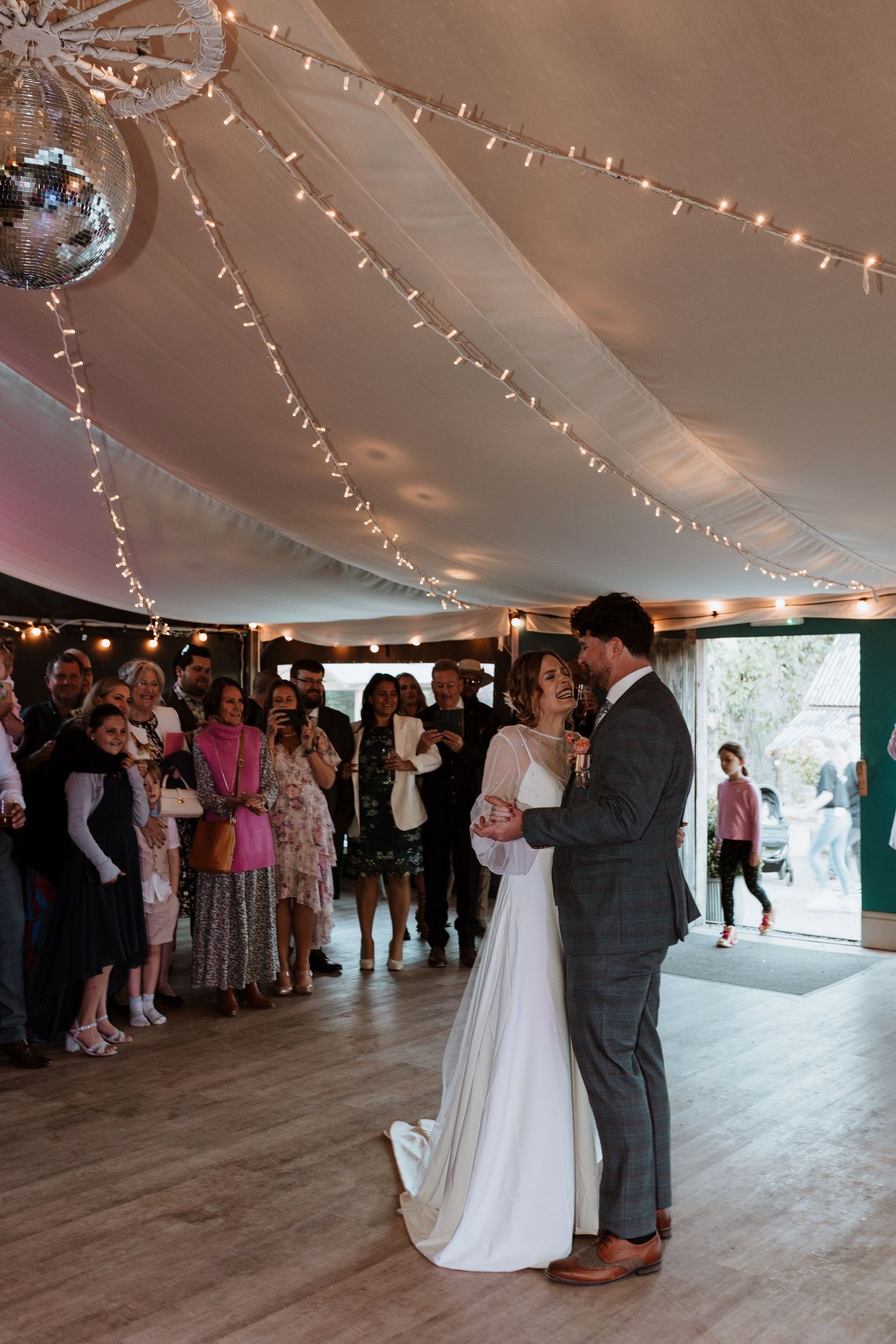 A couple shares their first dance under twinkling string lights while wedding guests look on in an elegant reception tent.