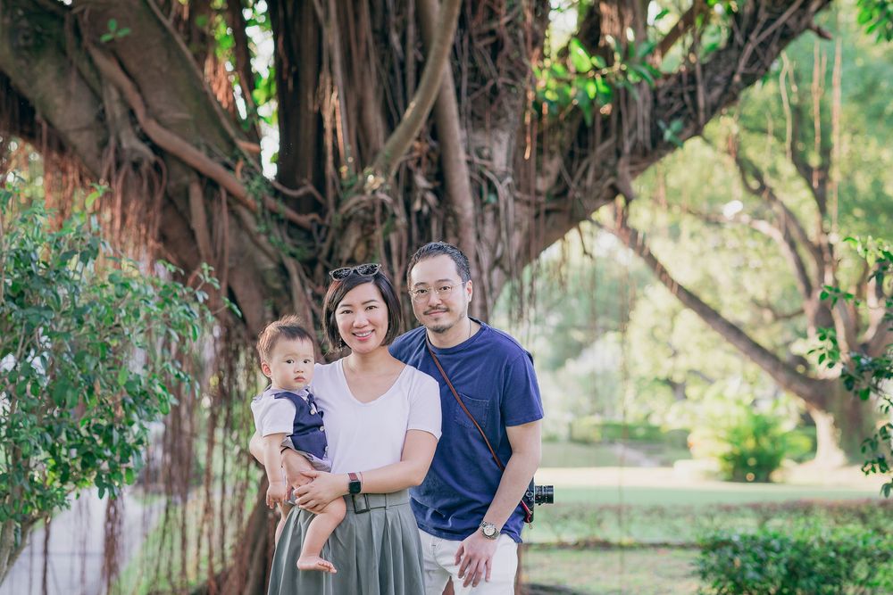 A family stands together smiling under a large banyan tree in a peaceful garden setting.