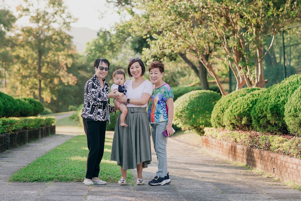 Three generations enjoy a sunny stroll together on a winding garden path lined with manicured shrubs and trees.