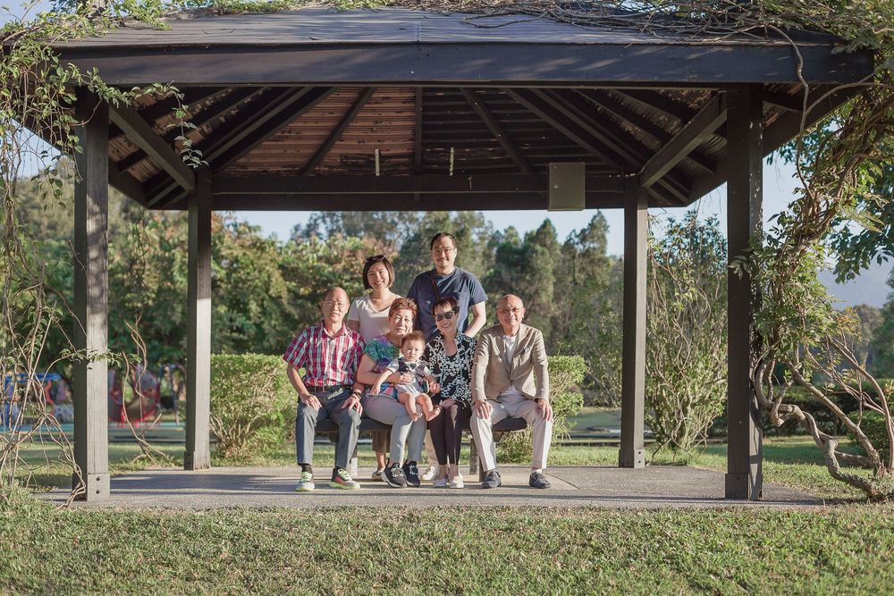 A group sits together under a wooden gazebo on a sunny day in a park-like setting.
