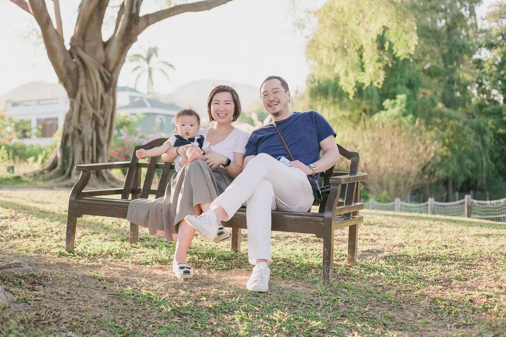 A family sits together on a wooden bench in a sunlit park with trees and greenery in the background.