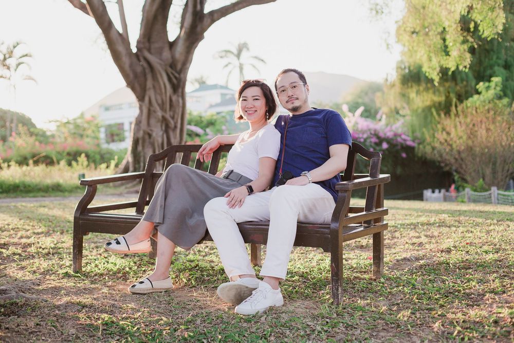 A couple relaxes together on a wooden bench in a sunny garden with trees and greenery in the background.