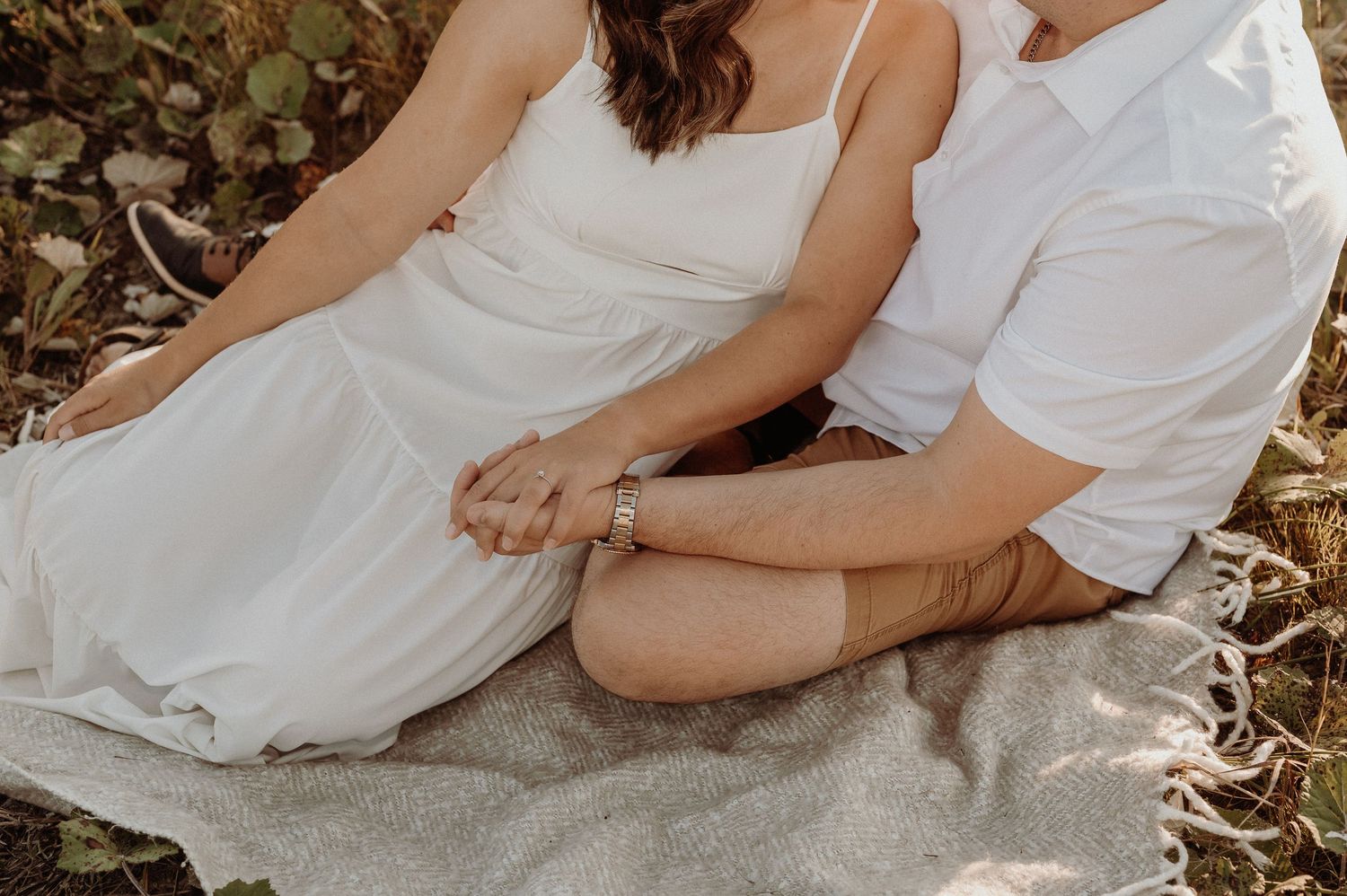 Couple wearing white outfits sits close together on gray blanket surrounded by autumn leaves.