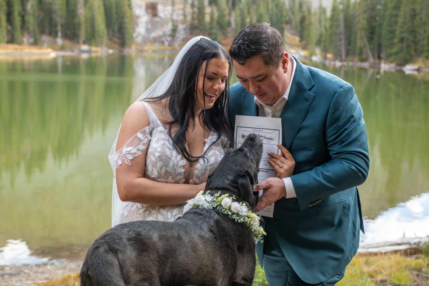 A bride and groom smile at their black dog who has just signed the marriage license they are holding. The dog has a white floral collar on.