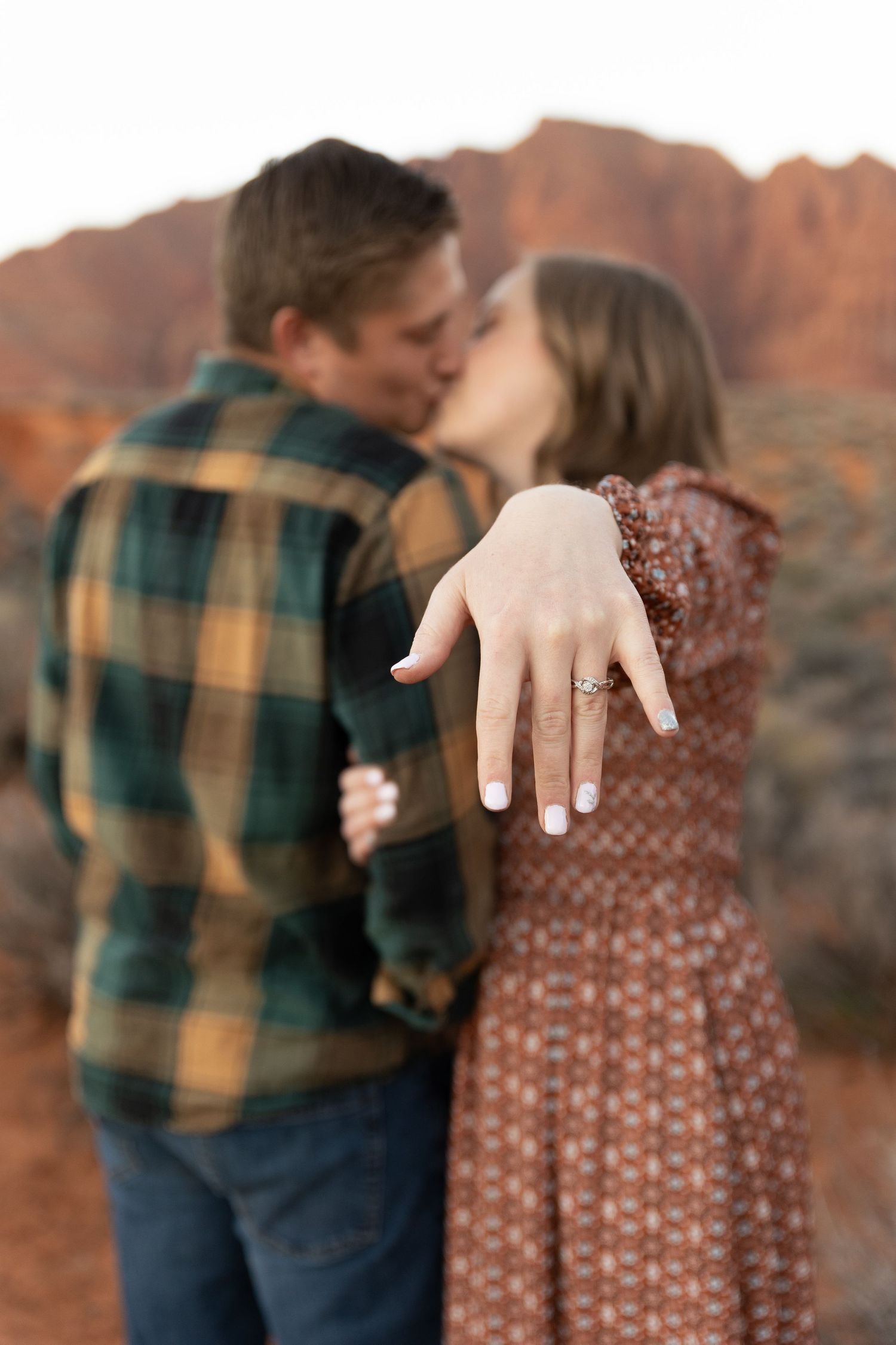 man and woman kissing blurred in the background with her hand outstreched showing off her ring