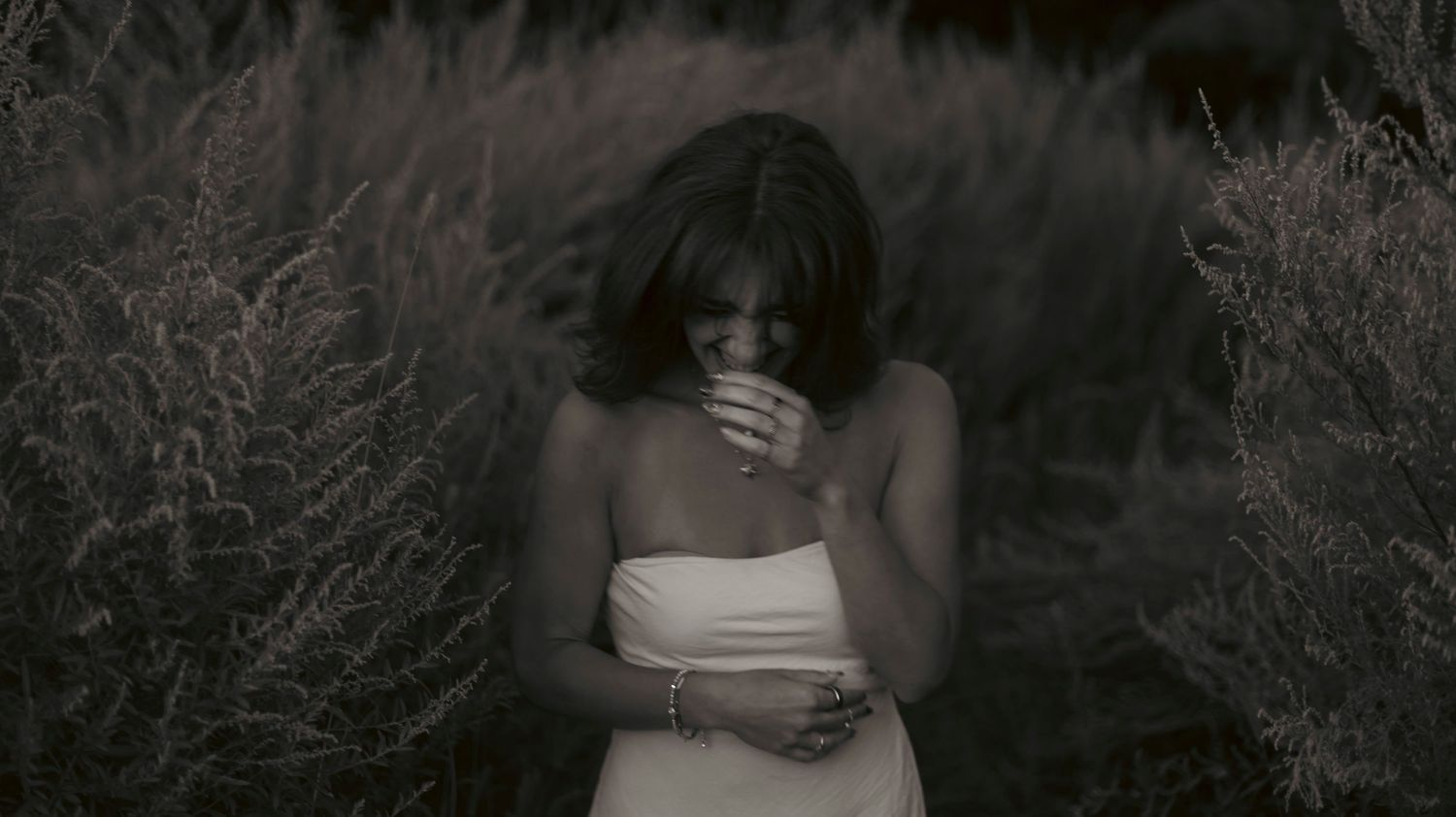 A person in a white top stands amid dark foliage while covering their face with their hand.