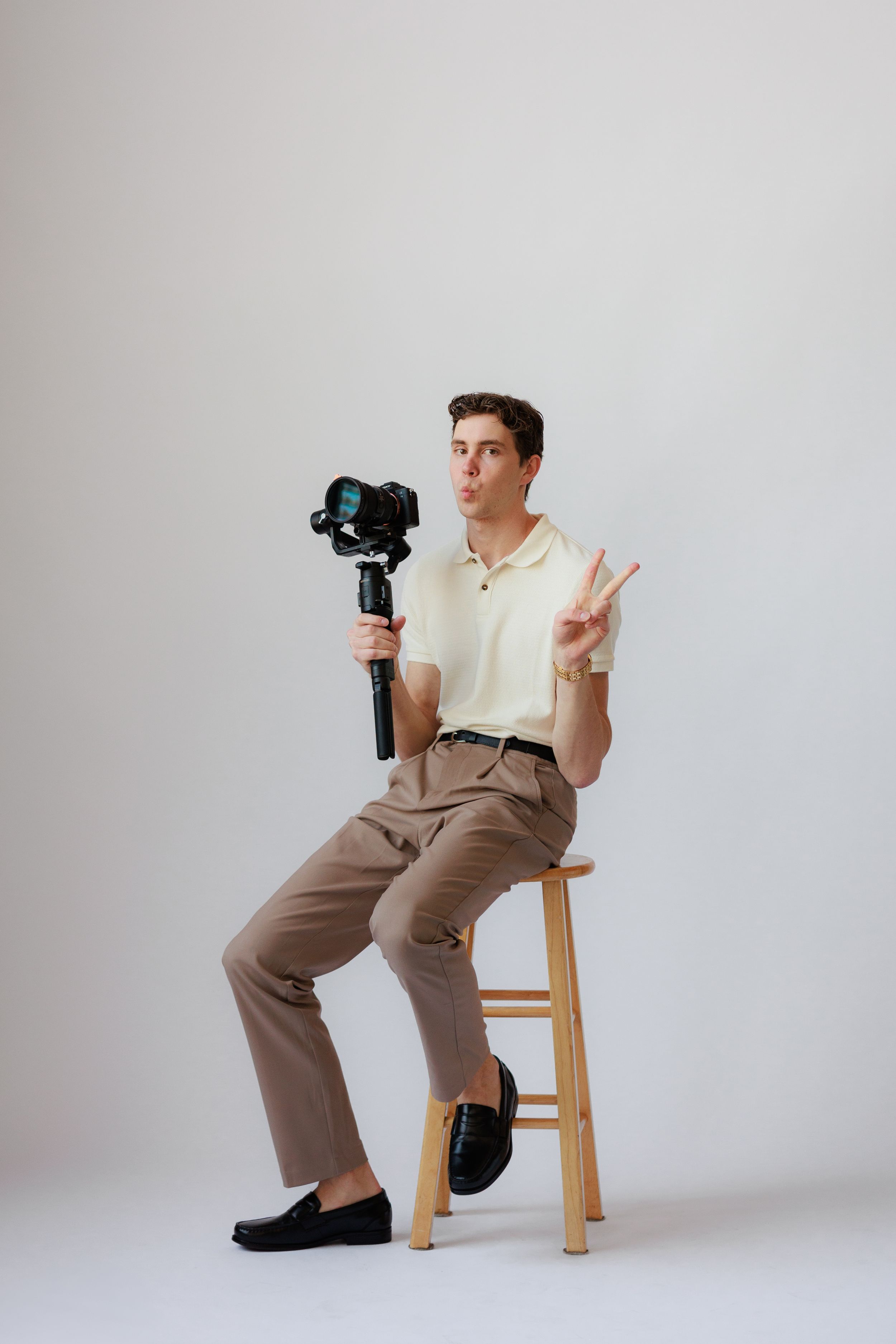 A person in khaki pants and light shirt sits on wooden stool making peace sign while holding camera in studio setting.