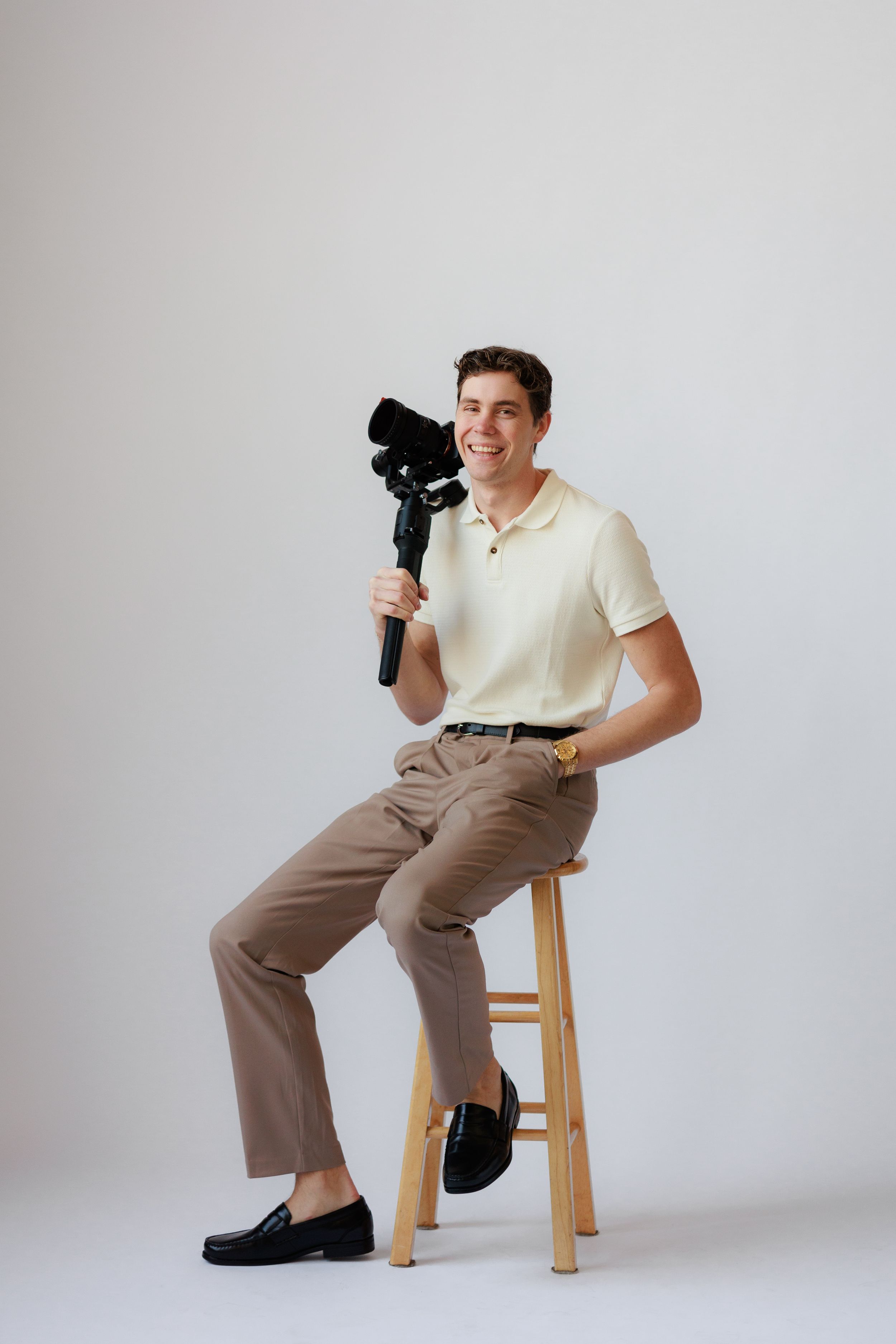 A photographer in a cream polo shirt and khaki pants sits on a wooden stool while holding a camera in a studio setting.