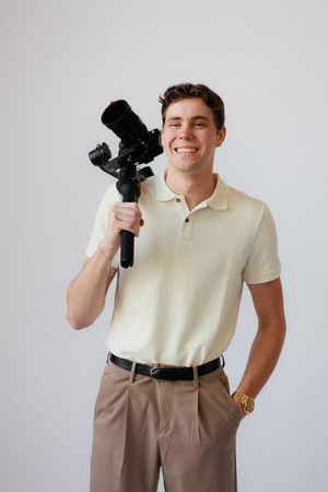 Person in cream polo shirt and brown pants poses playfully with camera equipment against white background.