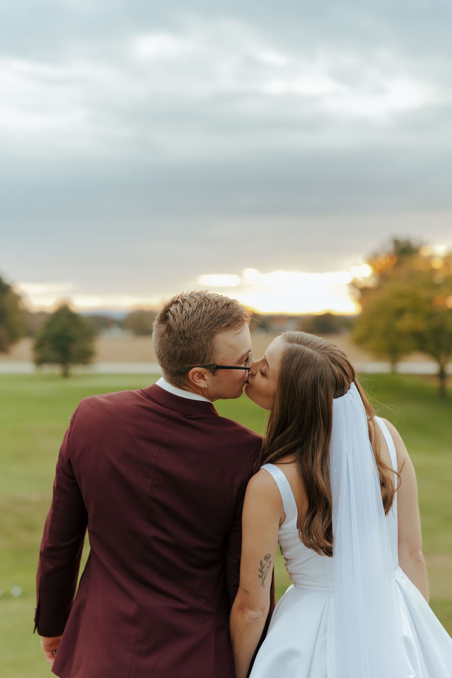 A couple in wedding attire share a romantic kiss at sunset on a golf course.