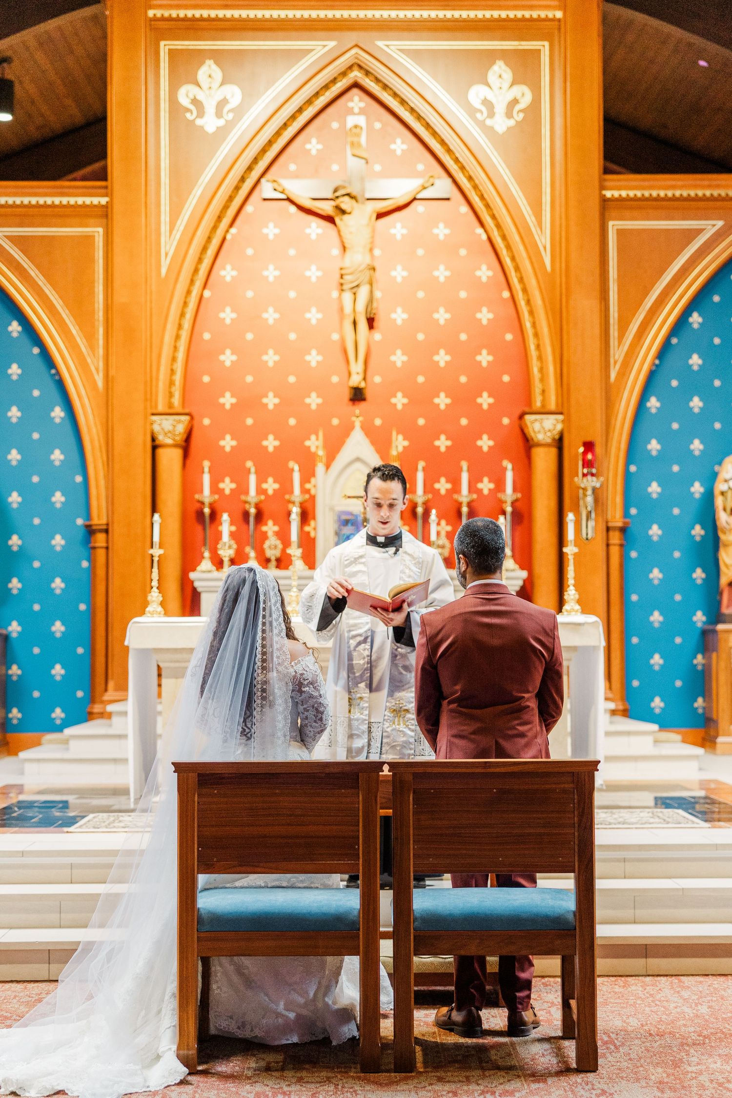 A wedding ceremony takes place in a Catholic church with ornate Gothic arches and stained glass windows in orange and blue tones.