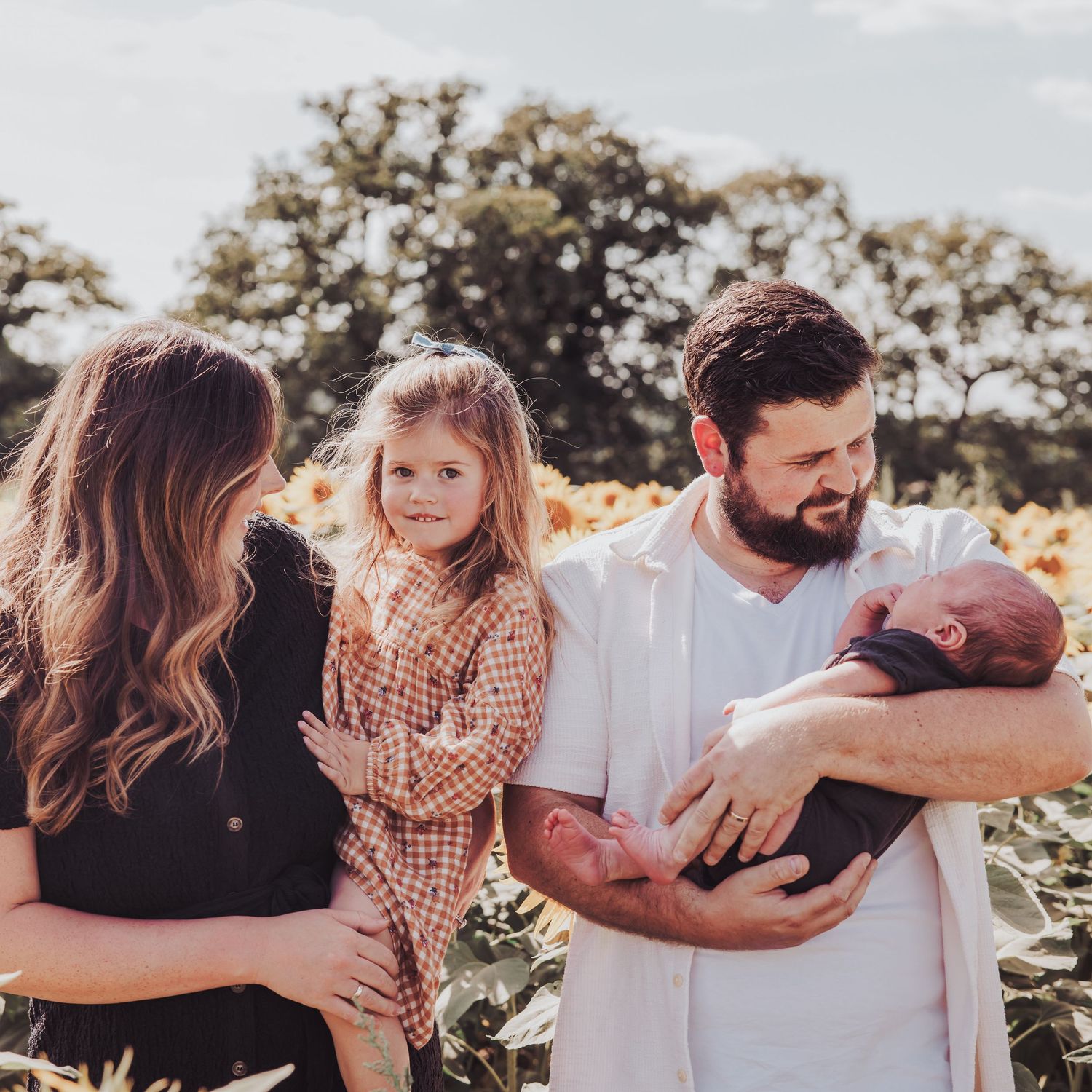 Parents and child standing together holding their newborn baby in a sunflower field.