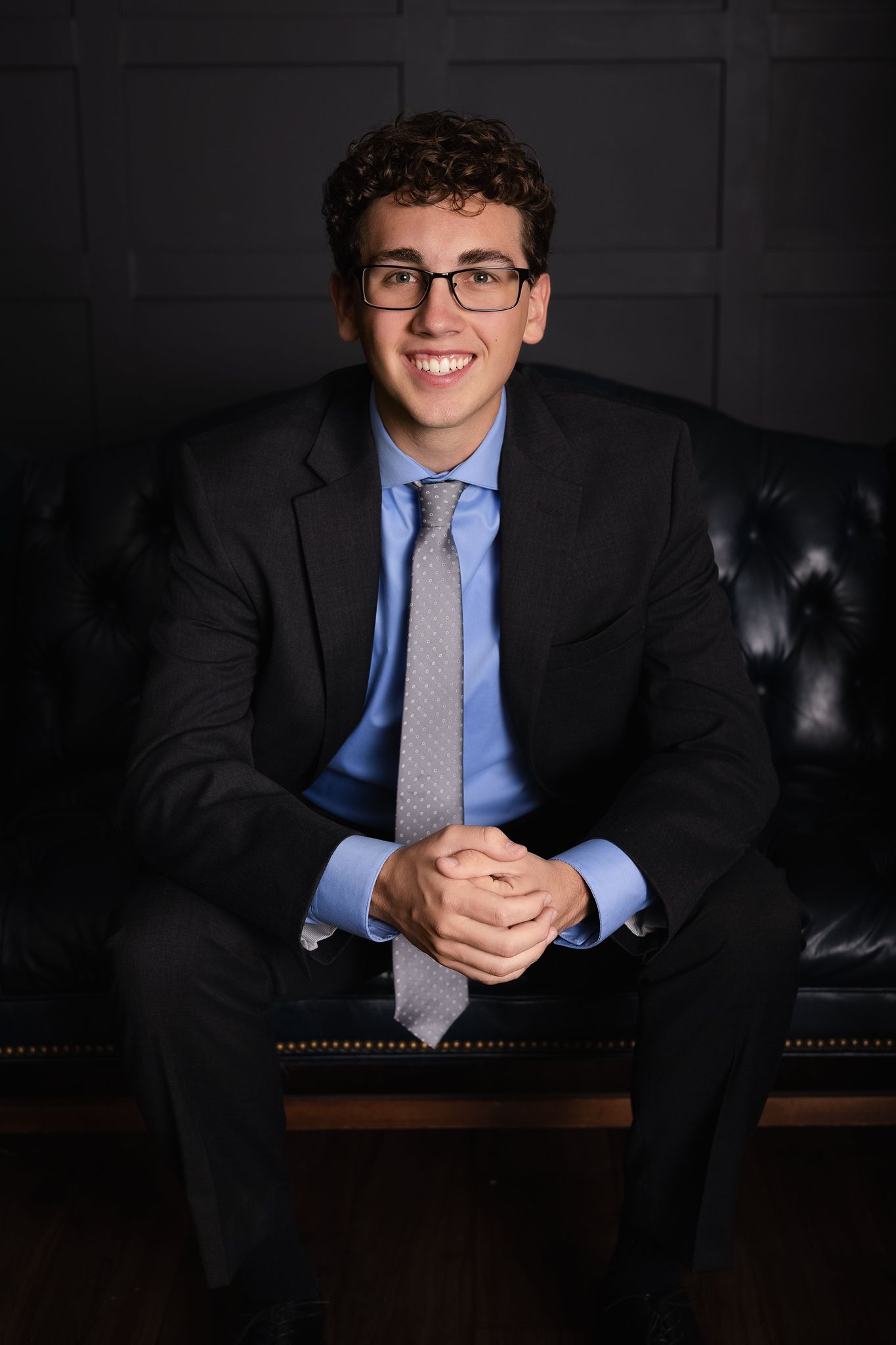 Professional headshot of someone in a dark suit and blue shirt with glasses seated on a leather couch in moody lighting.
