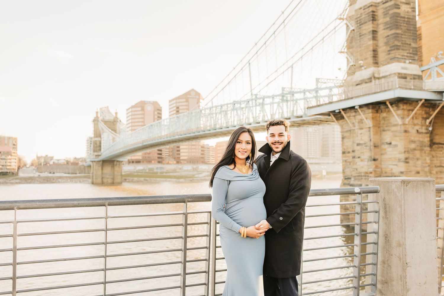 Couple embracing during a winter maternity photo shoot in downtown Cincinnati with Roebling Bridge in the background