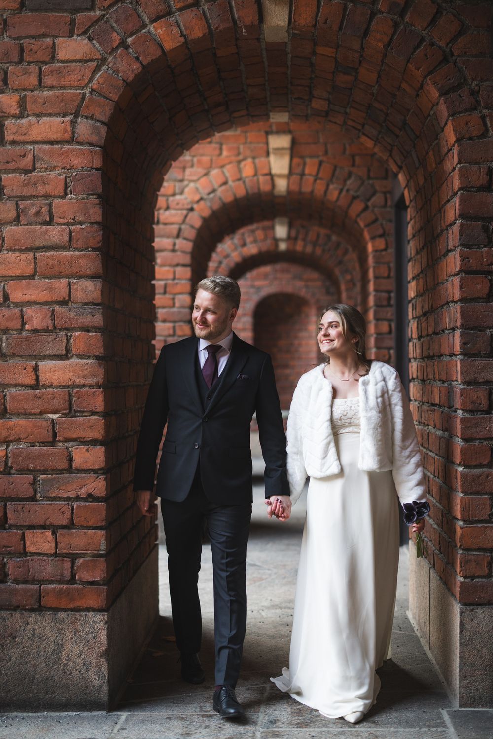 A couple stands in a romantic brick archway corridor, with the bride wearing a white dress and jacket.
