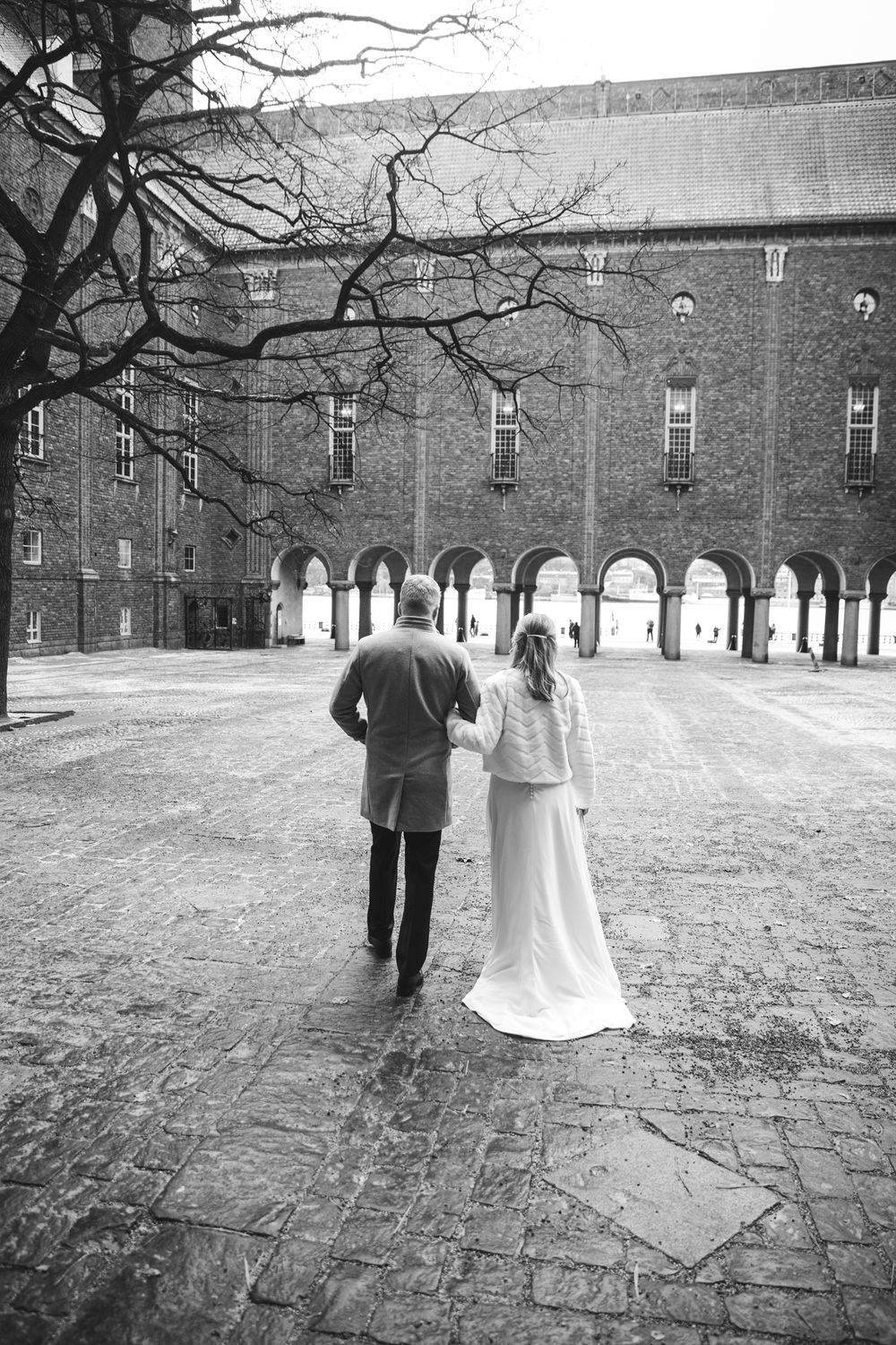 A wedding couple walks away from the camera across a stone courtyard with historic brick buildings in the background.