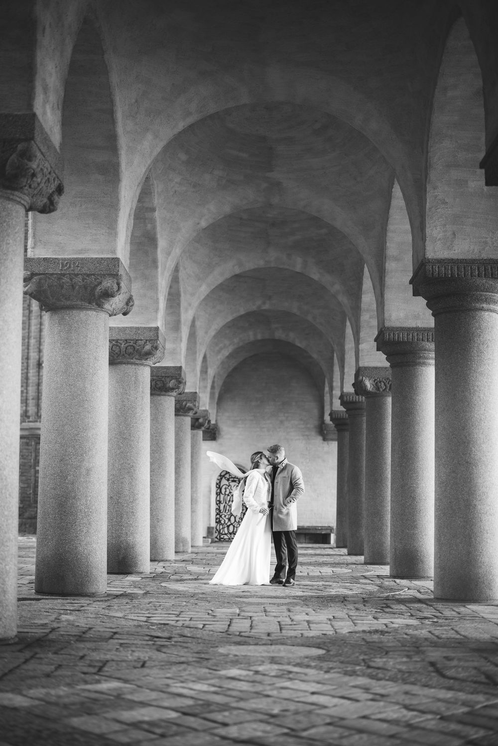 Dramatic black and white panoramic view of a stone colonnade with arched ceilings and a figure in the distance.