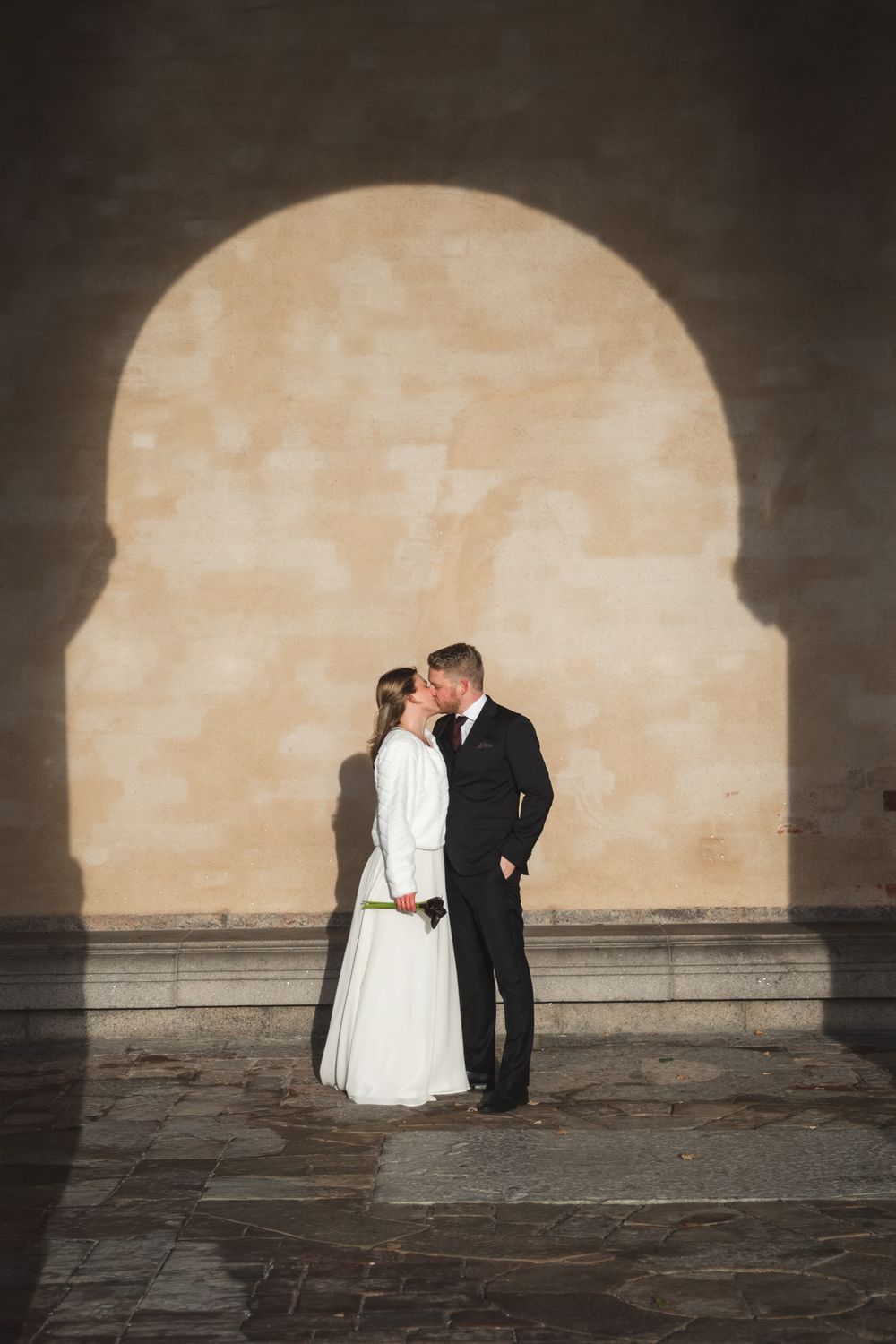 A romantic wedding scene unfolds in silhouette against a beige arched wall with stone flooring.