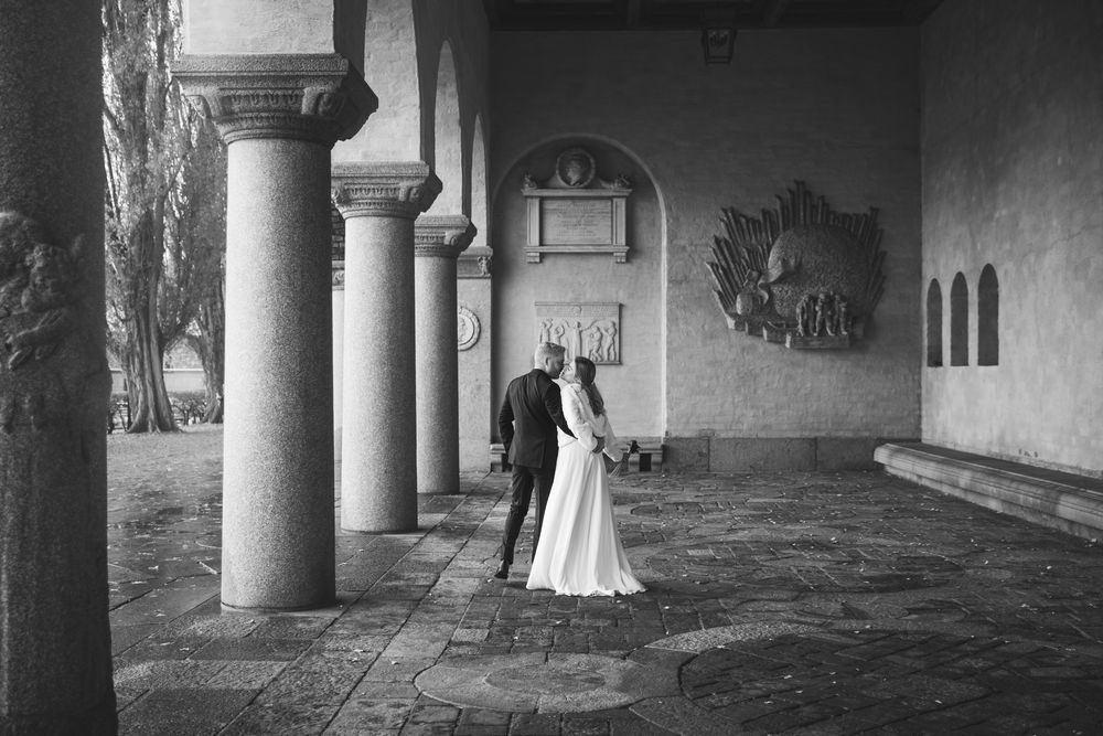 A black and white photograph shows a person in a long white dress walking through a columned corridor.