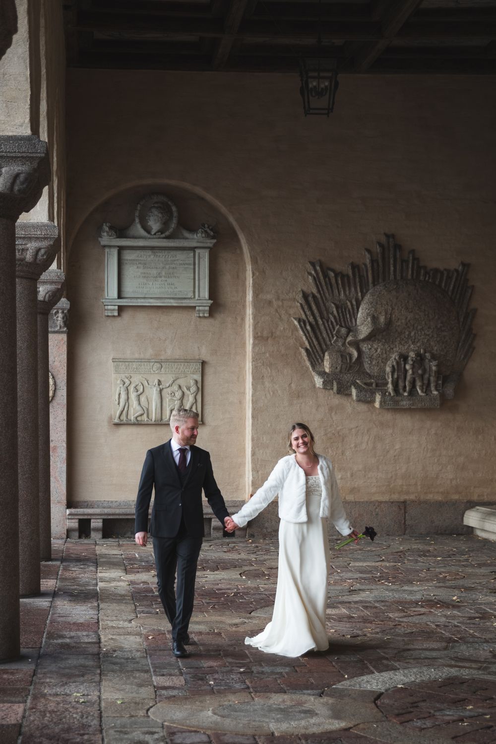 Wedding couple dances playfully in an elegant stone corridor with decorative wall art and brick flooring.