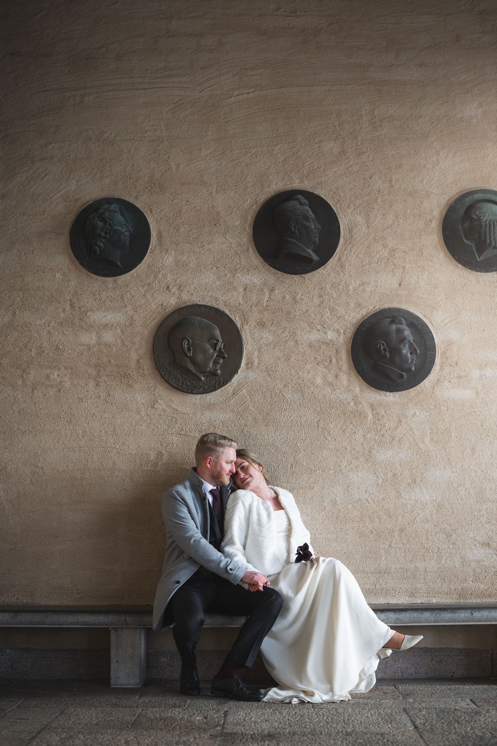 A couple in wedding attire sits together on a stone bench against a textured concrete wall with oval decorative elements.