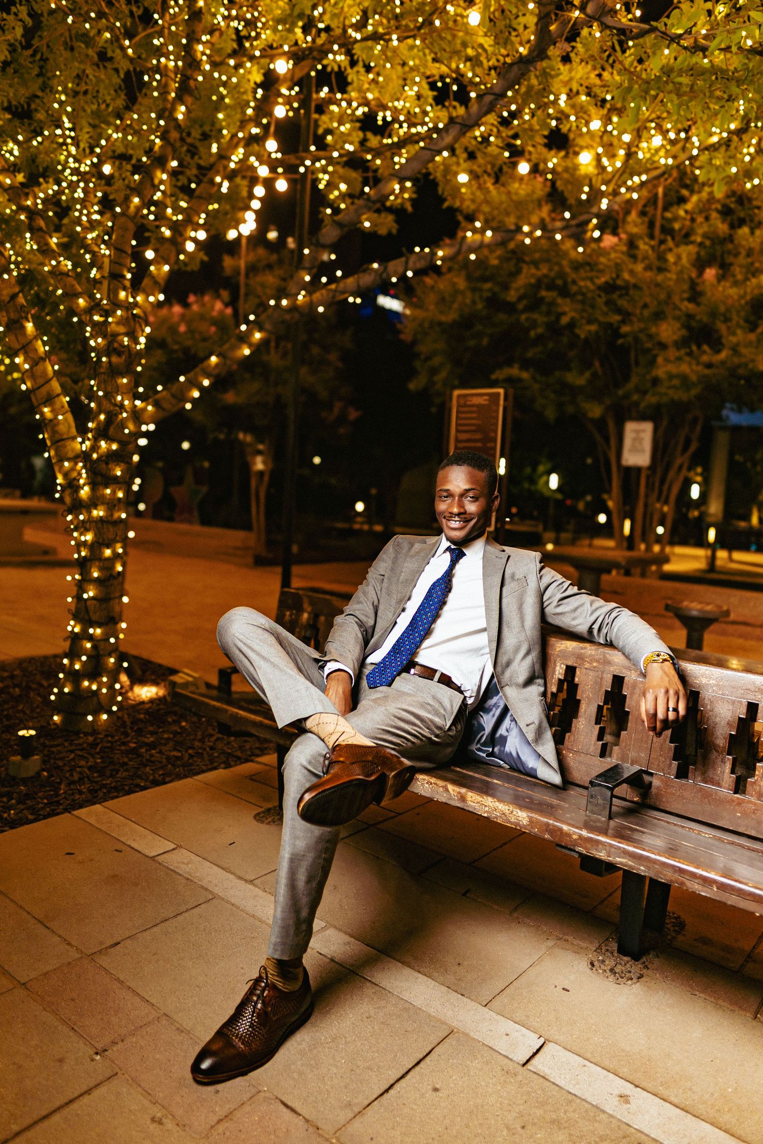 A person in a gray suit and blue tie relaxes on a bench under illuminated trees at night.