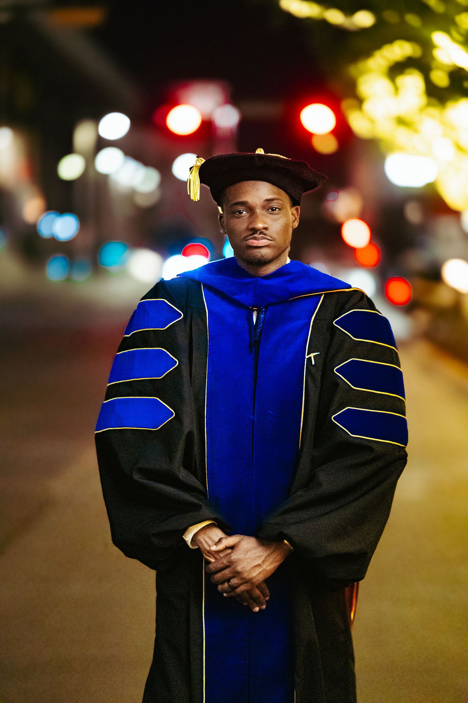 Graduate in academic regalia wearing a blue and black doctoral gown stands outdoors at night with city lights in background.