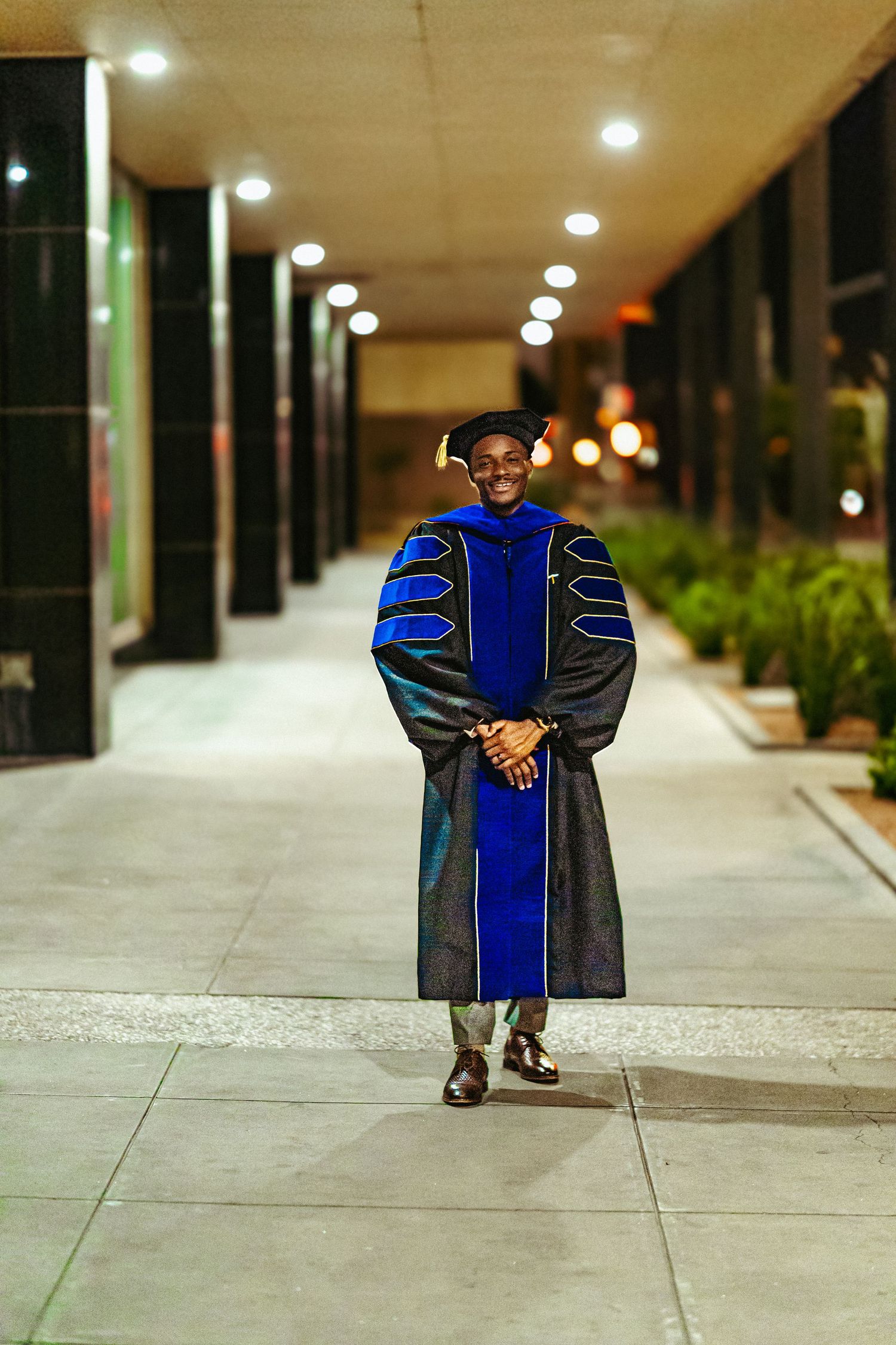 Graduate in doctoral regalia walks along illuminated walkway at night wearing blue and black academic gown.