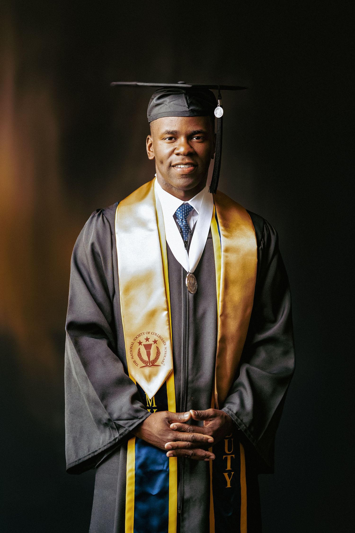 A graduate wearing black academic regalia with gold stole poses against a dark background for portraits.