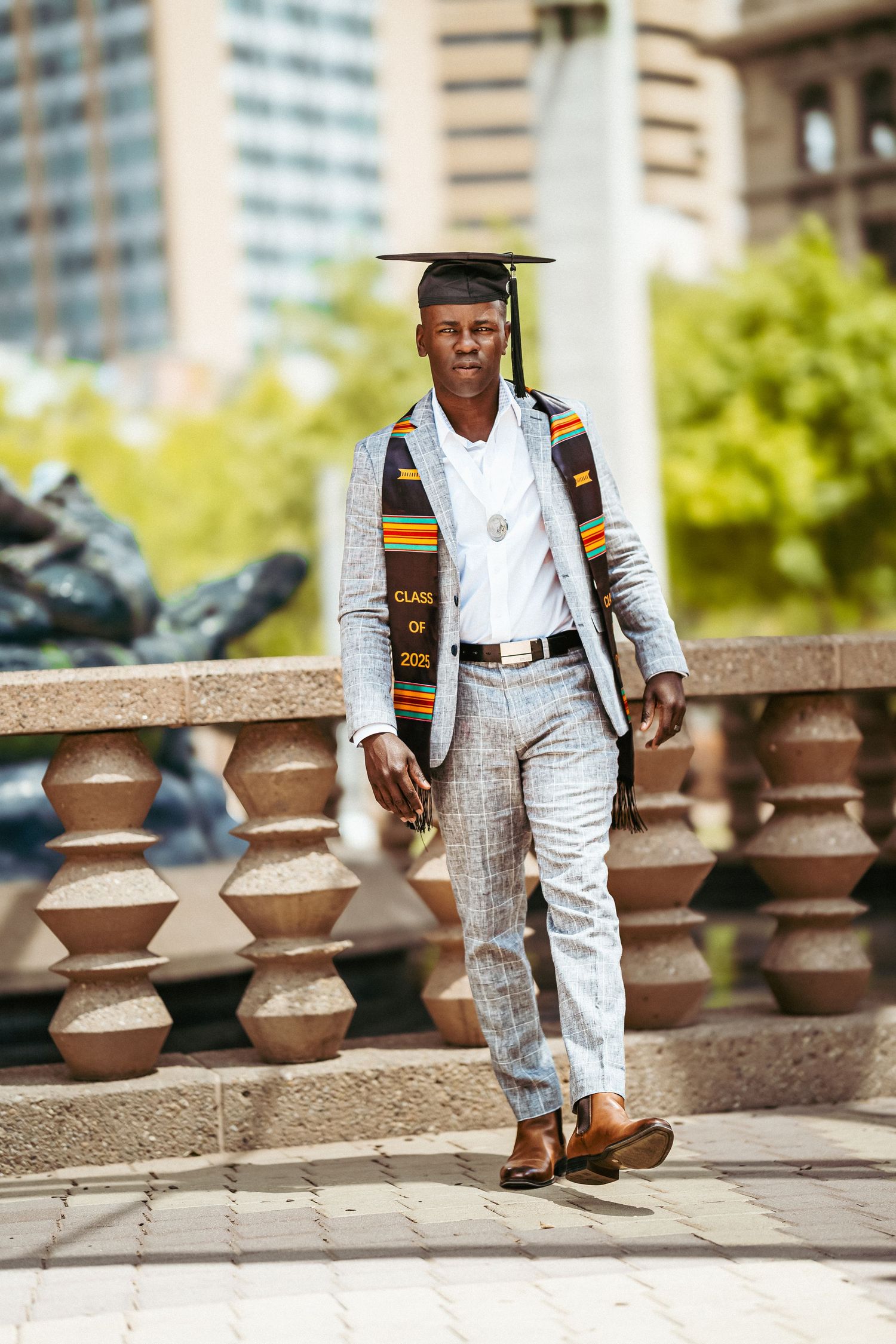 A graduate in a plaid suit and graduation cap poses confidently on stone steps near decorative concrete railings.