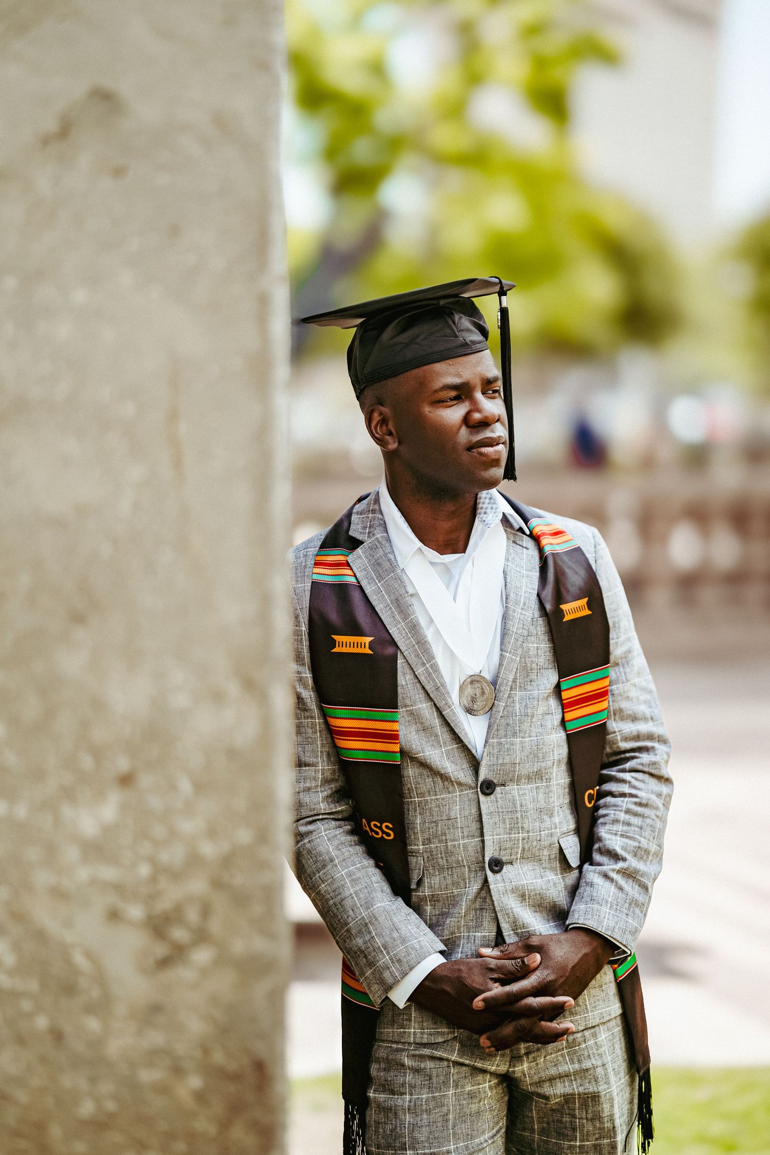 Graduate wearing traditional African stole and plaid suit poses for graduation photos outdoors on campus.